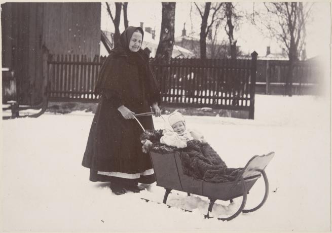 Foto: En bedstemor skubber et barn i en slæde ("isoäiti työntää lasta kelkassa"). Finnish Heritage Agency.