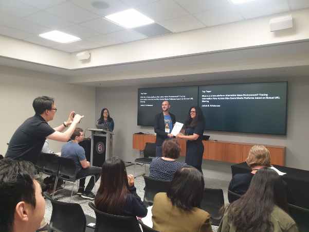 Picture of Jakob Bæk Kristensen receiving a best paper award. In front, seven people with their backs to the camera, one of them standing to the left to take a photo of the prize winner and the person who hands him the award. These two are facing the photo-taker. In the back a person standing behind a lectern.