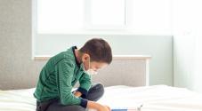 Child doing homework in bed wearing face mask