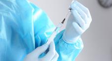 Person holding a surgical needle, wearing a blue surgeon's coat and plastic gloves
